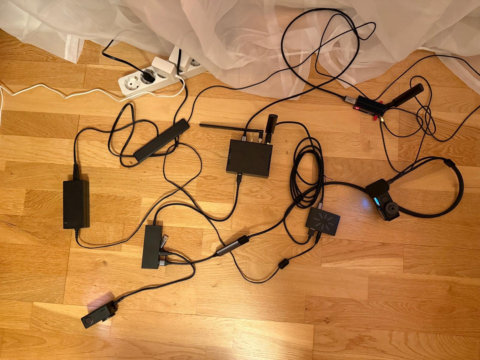 Bench prototype cabled up on a wooden floor: compute box with antennas, LTE modem on a USB M.2 adapter with heatsink, USB hub, capture dongle, power supply and two action cameras