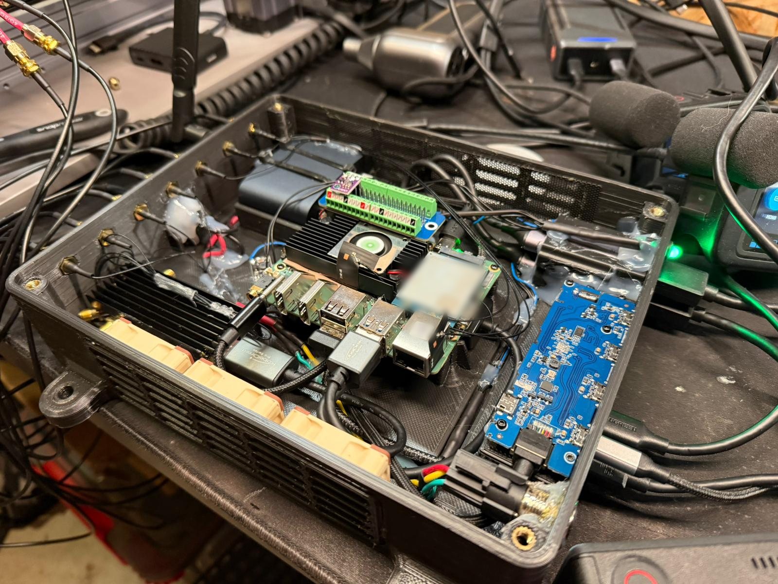Open 3D-printed testbed enclosure with a Radxa ROCK 5B+ under a fan cooler, an LTE modem, a green terminal board carrying a small sensor, a blue USB board, fans along the vented wall and SMA antenna pigtails